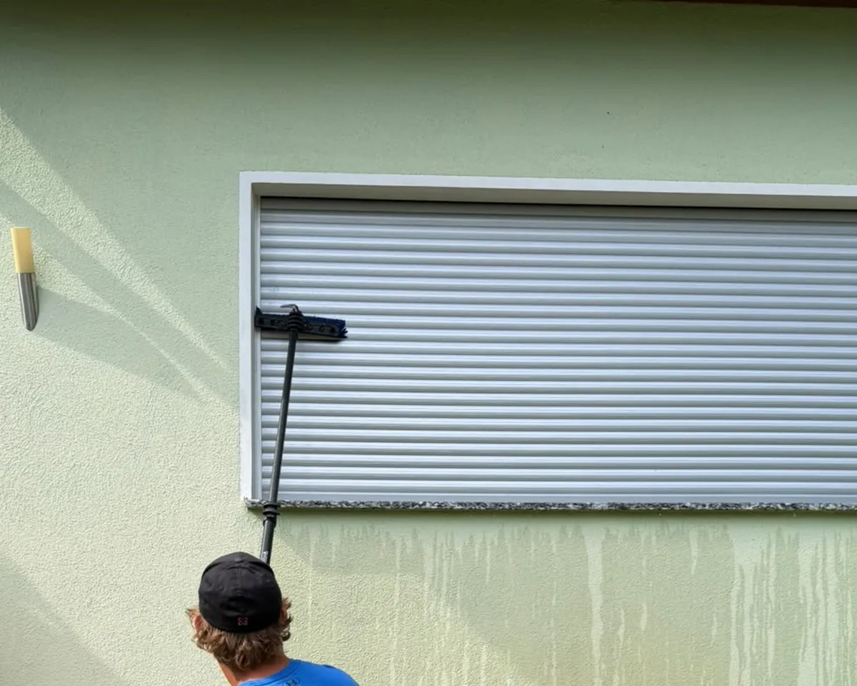 Person reinigt eine hellgrüne Putzfassade unterhalb eines Fensters mit einer Bürste an der Stange, auf der Wand nasse Reinigungsbahnen