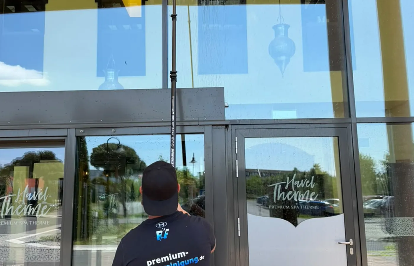 Mitarbeiter reinigt die hohe Eingangsverglasung einer Therme mit der wassergeführten Teleskopstange, auf den Glastüren der Schriftzug Havel-Therme