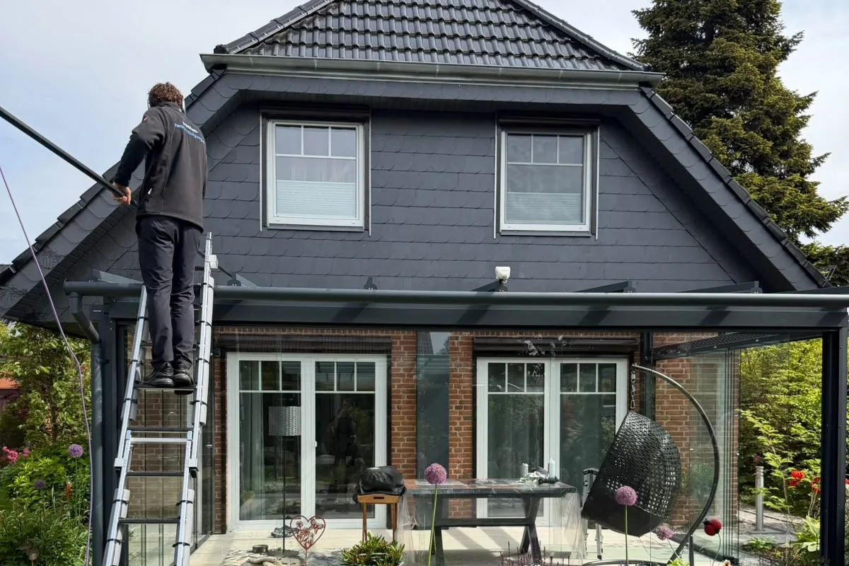 Mitarbeiter reinigt von einer Leiter aus das Glasdach eines Wintergartens mit einer Wasserstange, dahinter ein Backsteinhaus, davor Terrasse und Staudenbeet