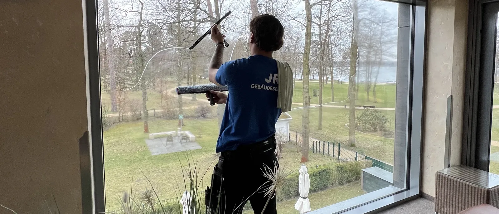 Mitarbeiter reinigt eine raumhohe Fensterfront von innen mit Einwascher und Abzieher, dahinter Garten und Seeufer